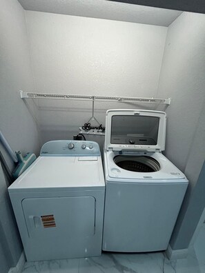 Ein kleiner Hauswirtschaftsraum mit einer Waschmaschine und einem Trockner mit Frontlader, einem Wäscheständer aus Metall und einem an der Wand befestigten Schild.