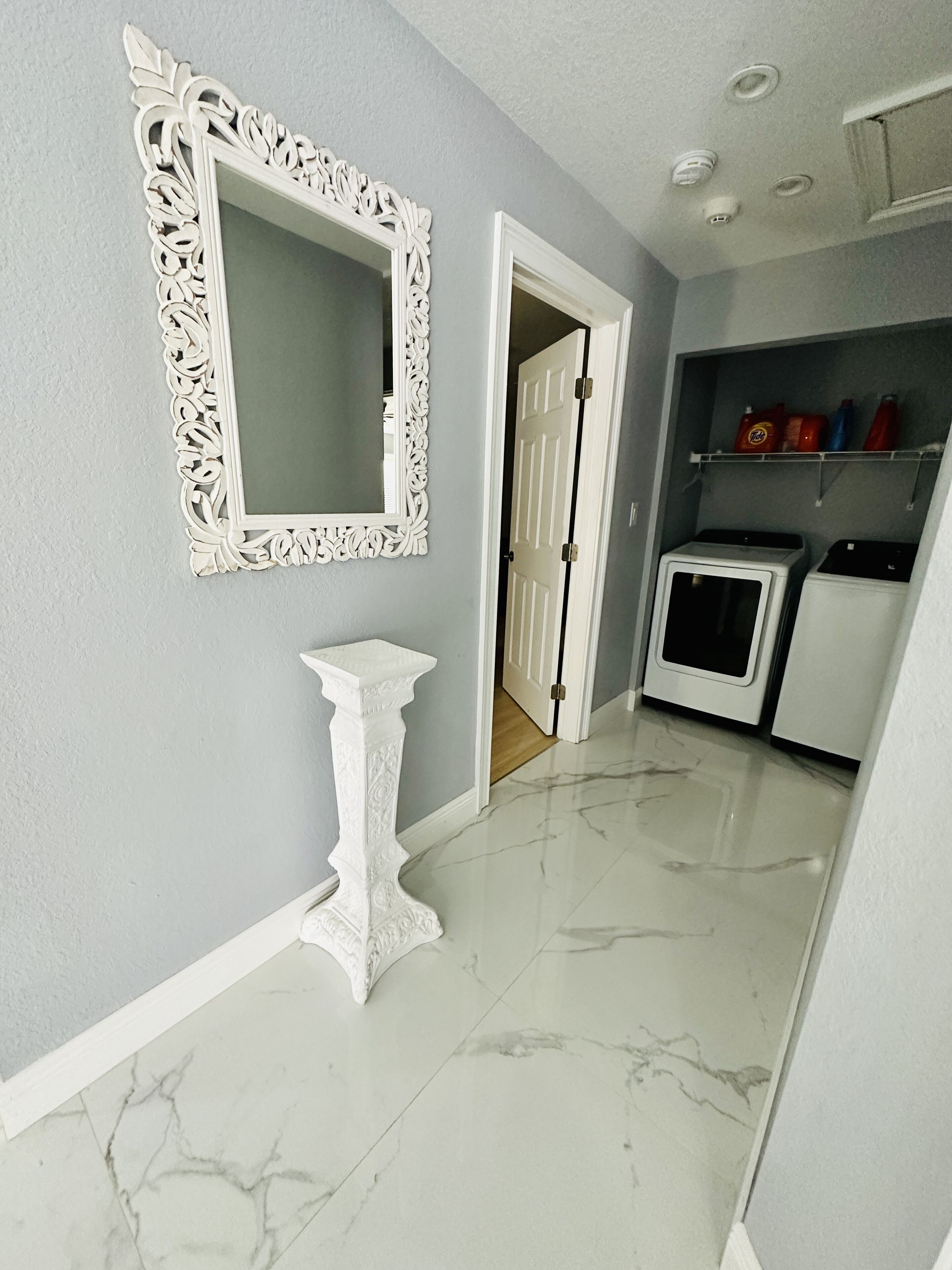 A hallway with a white ornate mirror, a pedestal table, and a laundry area with washing machines.