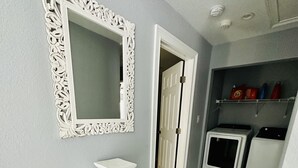 Un pasillo con un espejo blanco ornamentado, una mesita y un área de lavandería con lavadoras.