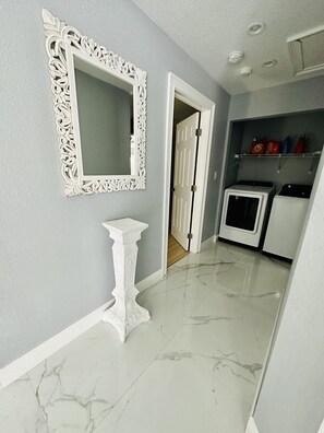 Un pasillo con un espejo blanco ornamentado, una mesita y un área de lavandería con lavadoras.