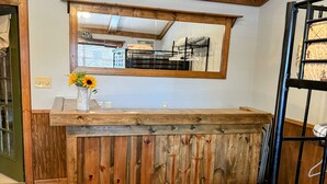 Uma área rústica com balcão de madeira e um espelho acima, um vaso com flores e uma cama beliche ao fundo.
