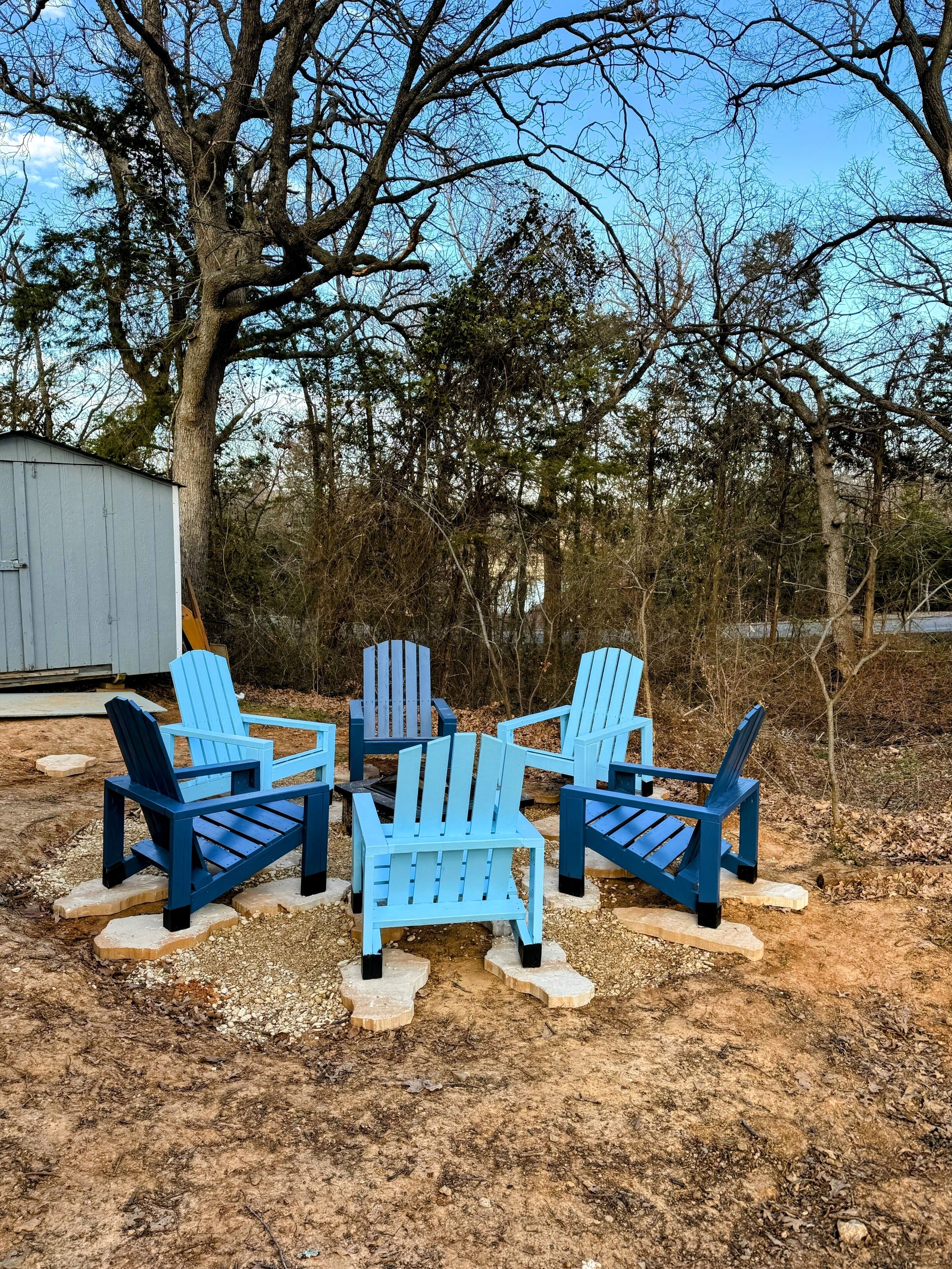 A set of four blue Adirondack chairs arranged around a small fire pit in a wooded area with a shed in the background.