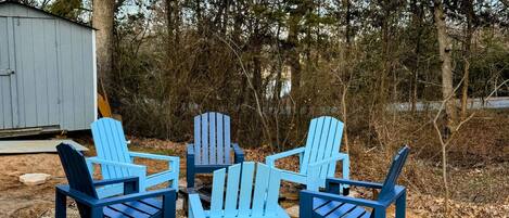 A set of four blue Adirondack chairs arranged around a small fire pit in a wooded area with a shed in the background.