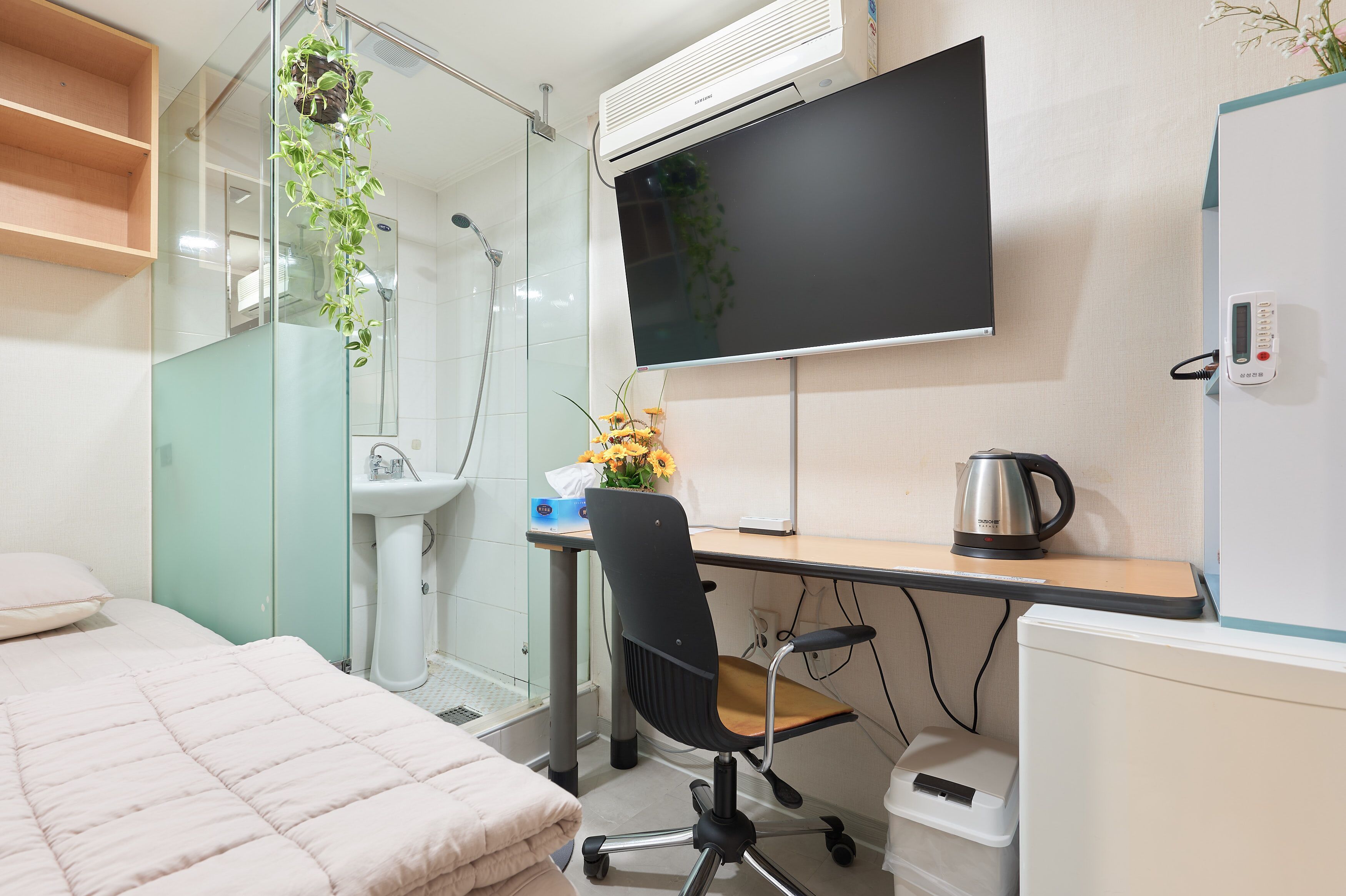 A compact hotel room with a bed, a desk with a chair, a TV, a sink, and a shower.