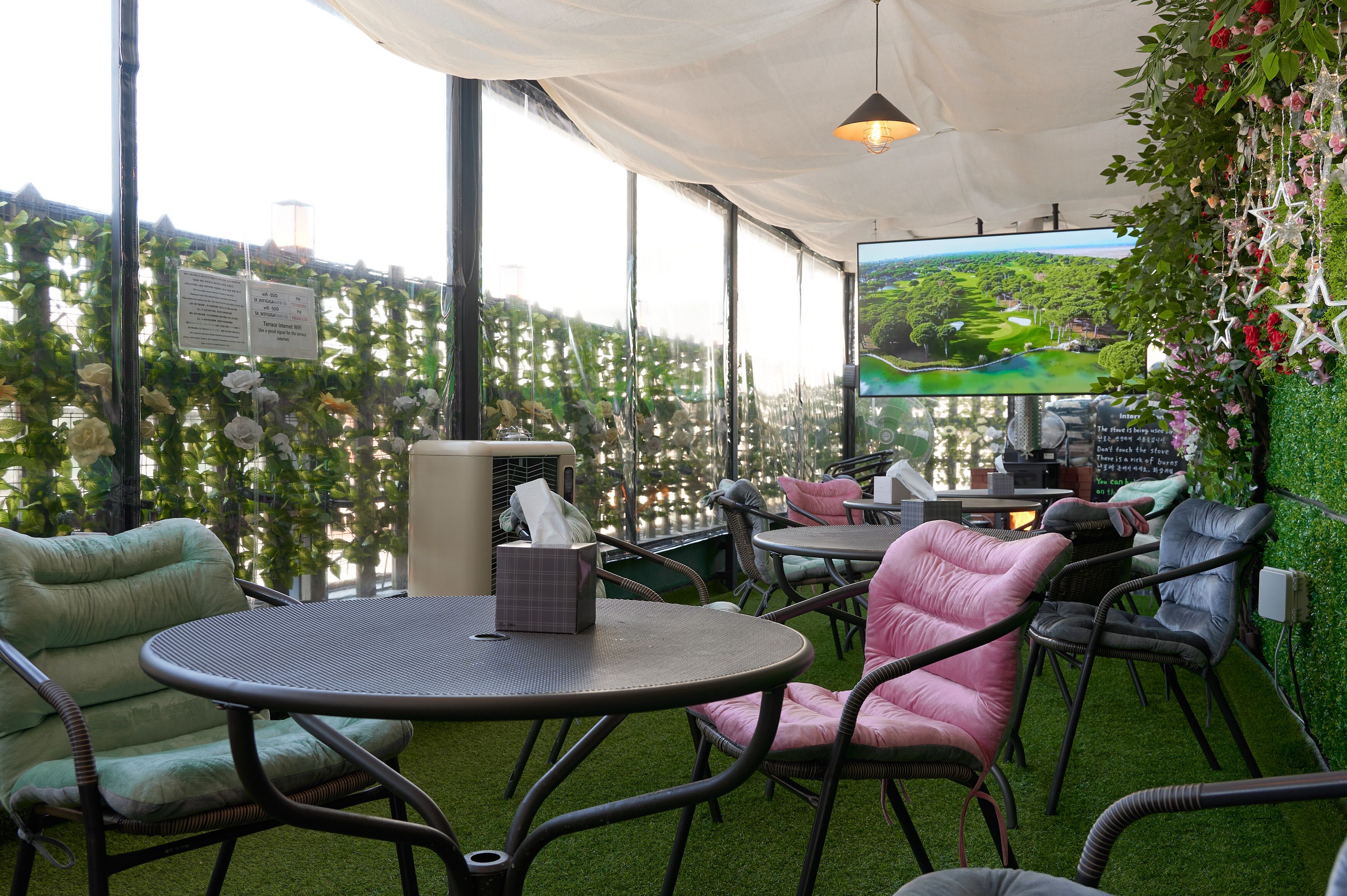 An outdoor seating area with round tables and chairs, a large screen displaying a scenic view, and a wall decorated with greenery and flowers.