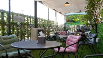 An outdoor seating area with round tables and chairs, a large screen displaying a scenic view, and a wall decorated with greenery and flowers.