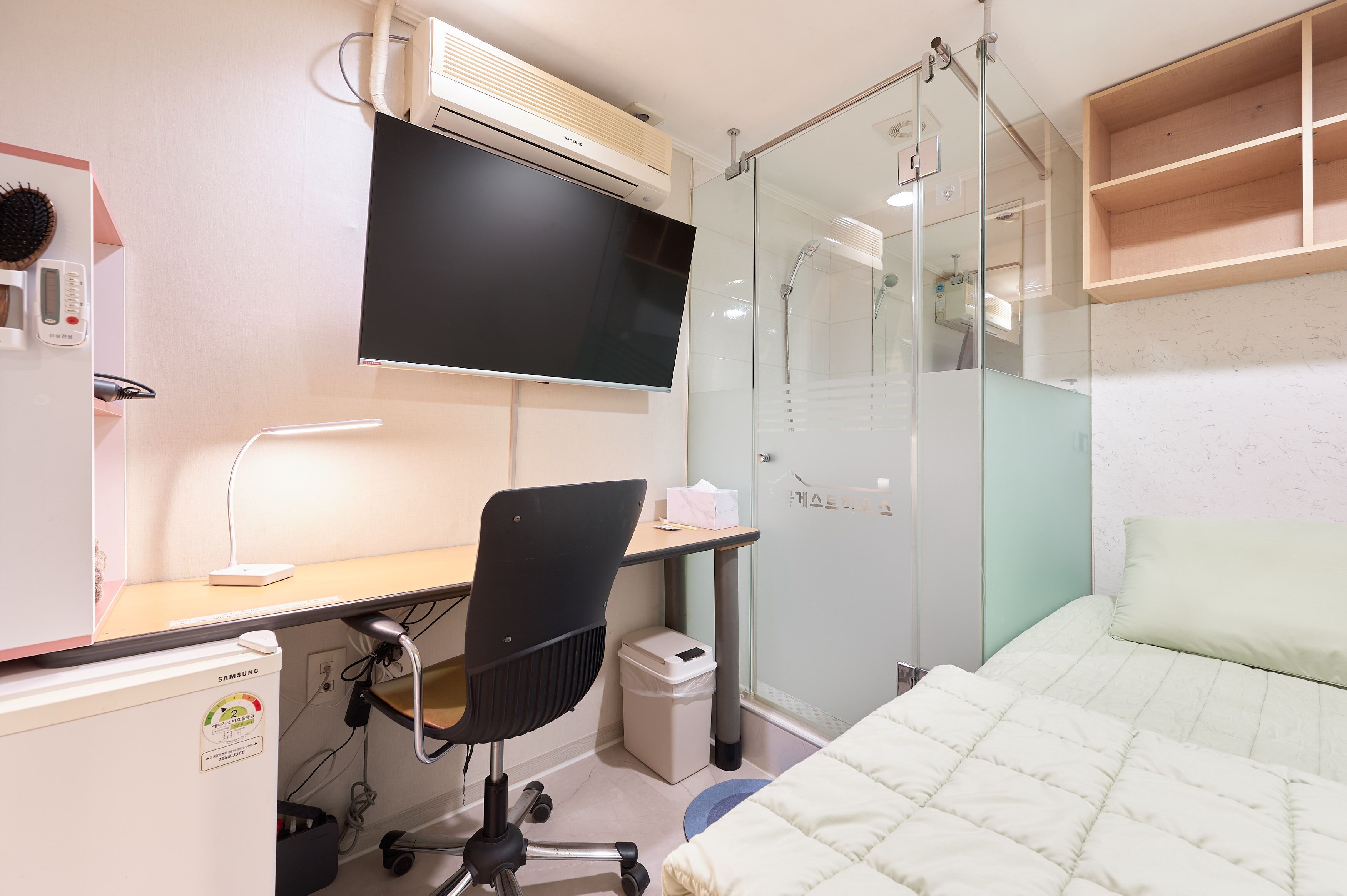 A hotel room with a bed, a desk with a chair, a TV, a shower, and a small trash bin.