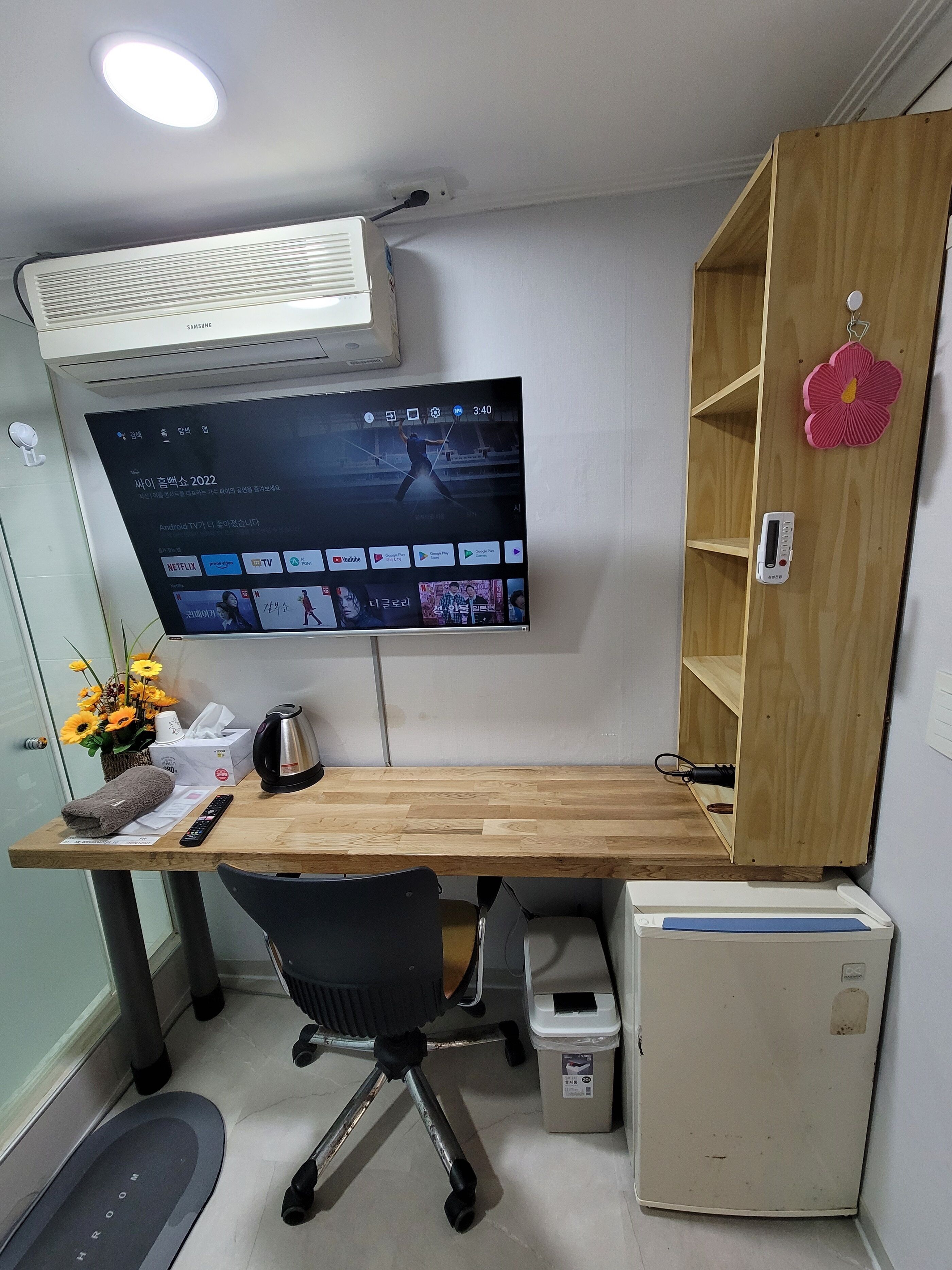 A compact office space with a wooden desk, a TV mounted on the wall, a small refrigerator, and a trash bin.