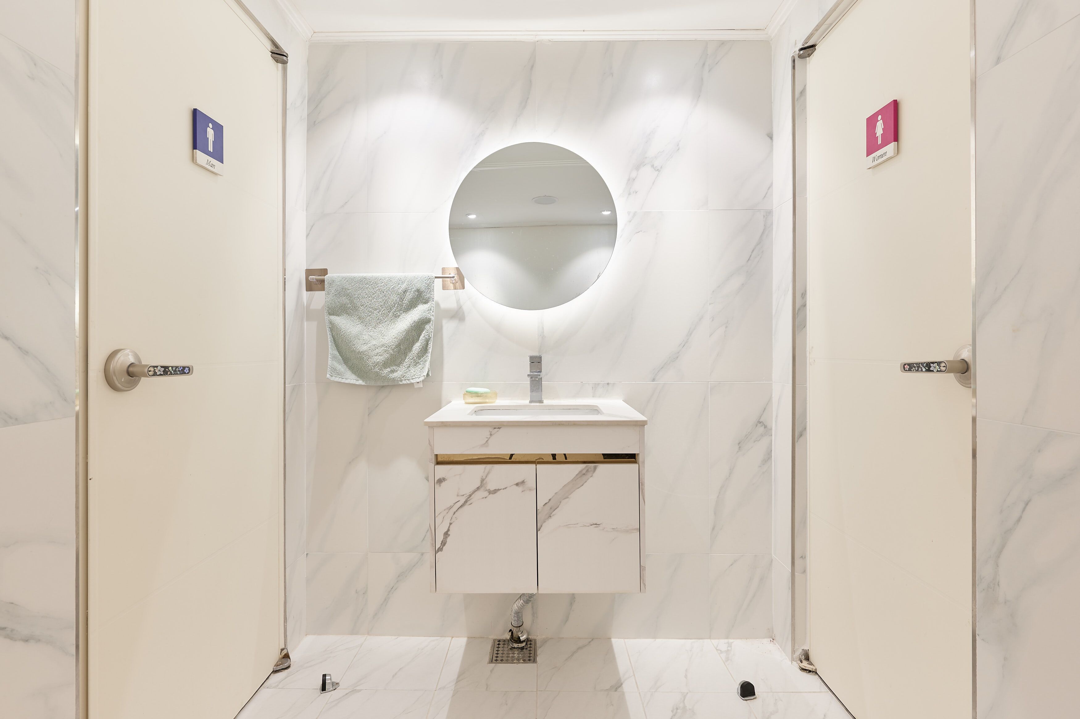 A bathroom with a marble vanity, a round mirror, and two doors with gender-specific signs.