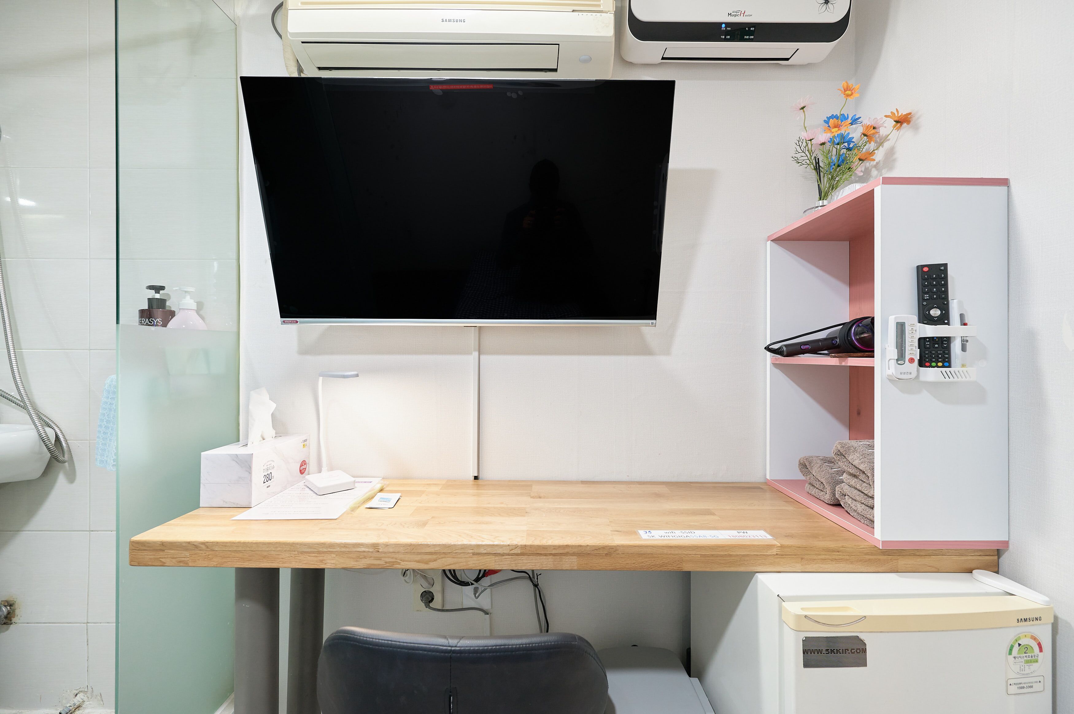 A hotel room with a wooden desk, a flat-screen TV mounted on the wall, a built-in air conditioning unit, a shelf with towels, and a small refrigerator.