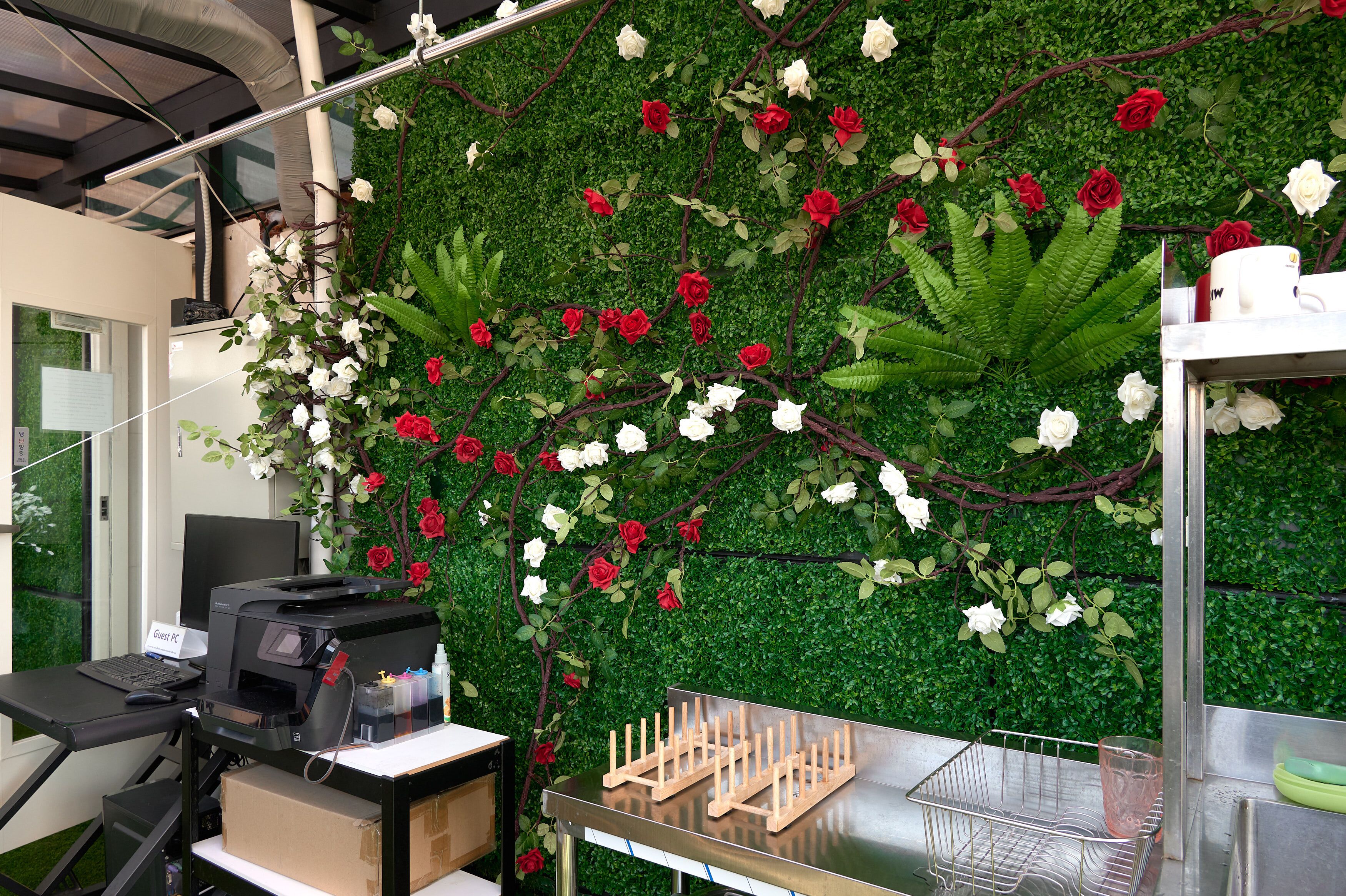 Un espacio de trabajo con una pared verde decorada con flores rojas y blancas, una impresora y una mesa metálica con un recipiente de vidrio.