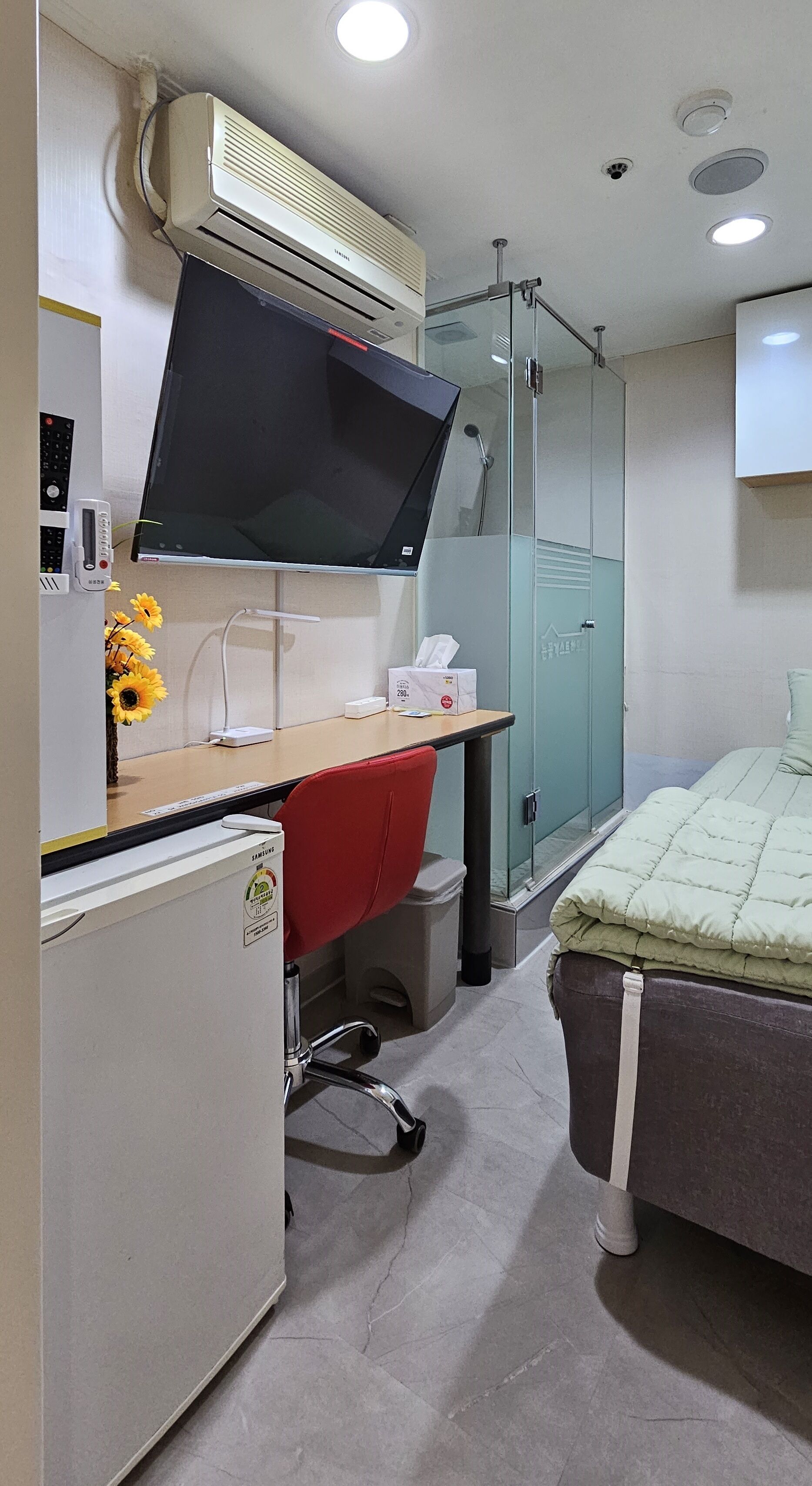 A compact hotel room with a desk, a bed, a shower, and a built-in air conditioner.