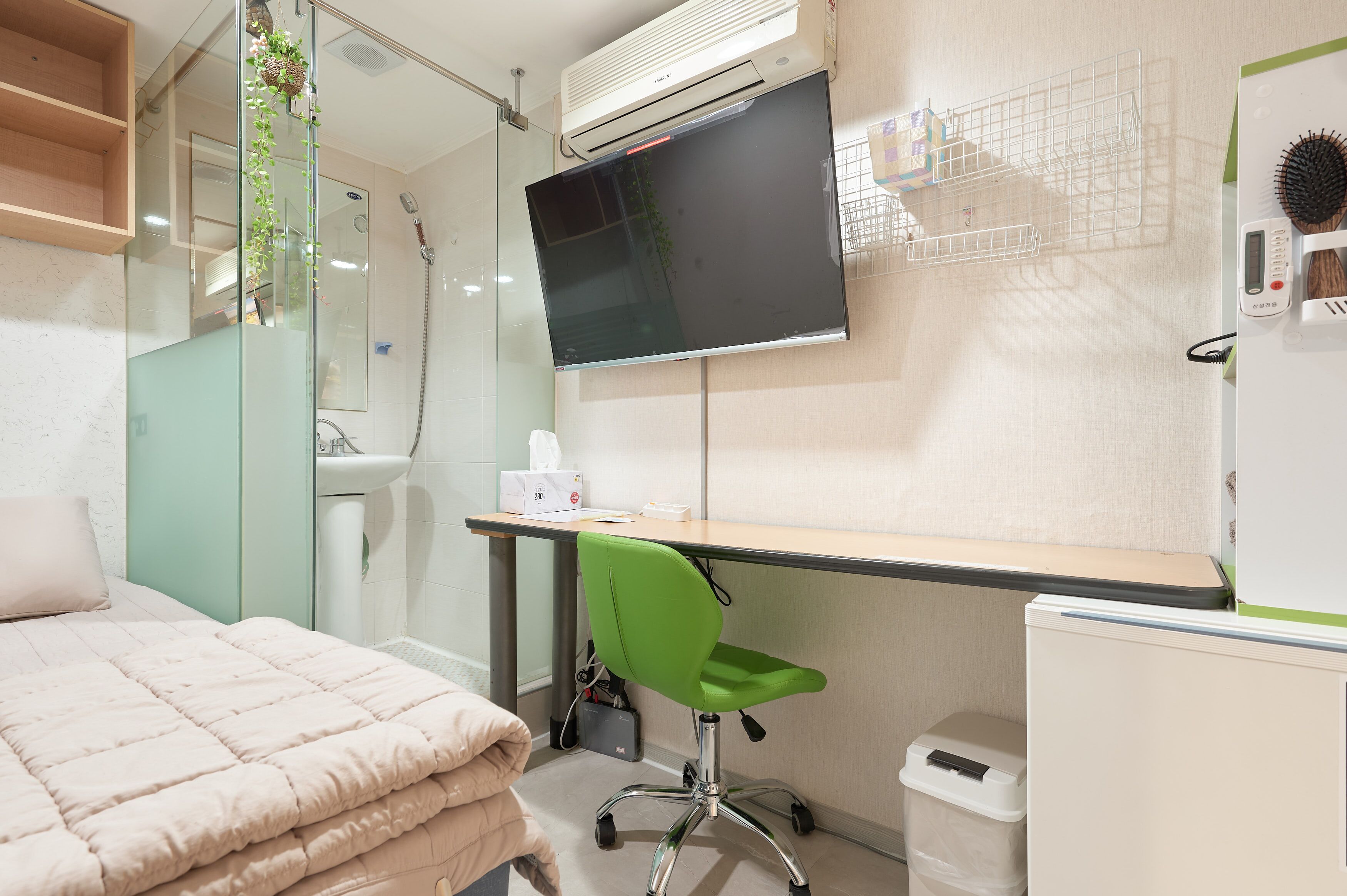 Een moderne slaapkamer met een bed, een bureau met een computer, een groene stoel, een televisie en een kleine prullenbak.