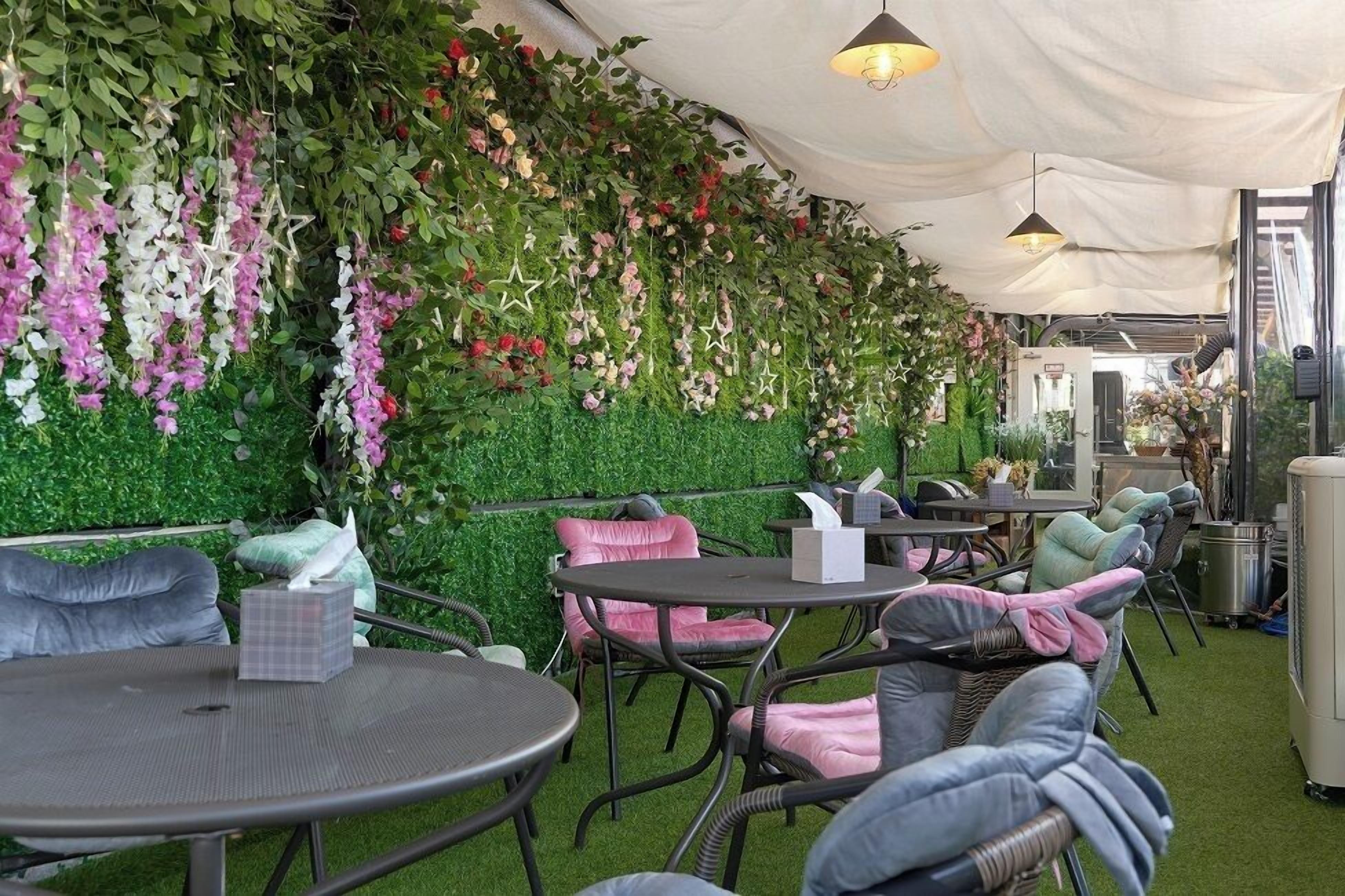 An indoor patio with round tables and cushioned chairs, surrounded by green foliage and hanging floral arrangements.