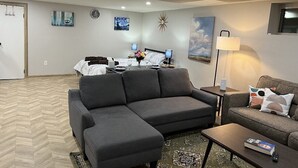 Un soggiorno moderno con un divano componibile, un tavolino e una TV. Si intravede un letto con un comodino e una lampada sullo sfondo.