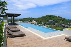 A rooftop infinity pool with wooden deck and lounge chairs, overlooking a coastal town with buildings and greenery. - Deluxe apartment at Panora by Lofty (Choeng Thale)