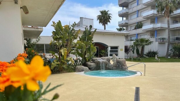 A pool area with a water feature, surrounded by tropical plants and palm trees, with a building in the background. - Welcome to the charming apartment for rent in Roses, Costa Brava! 1500 meters from the beach, modern accommodation with canal views, sleeps 4. (Roses)