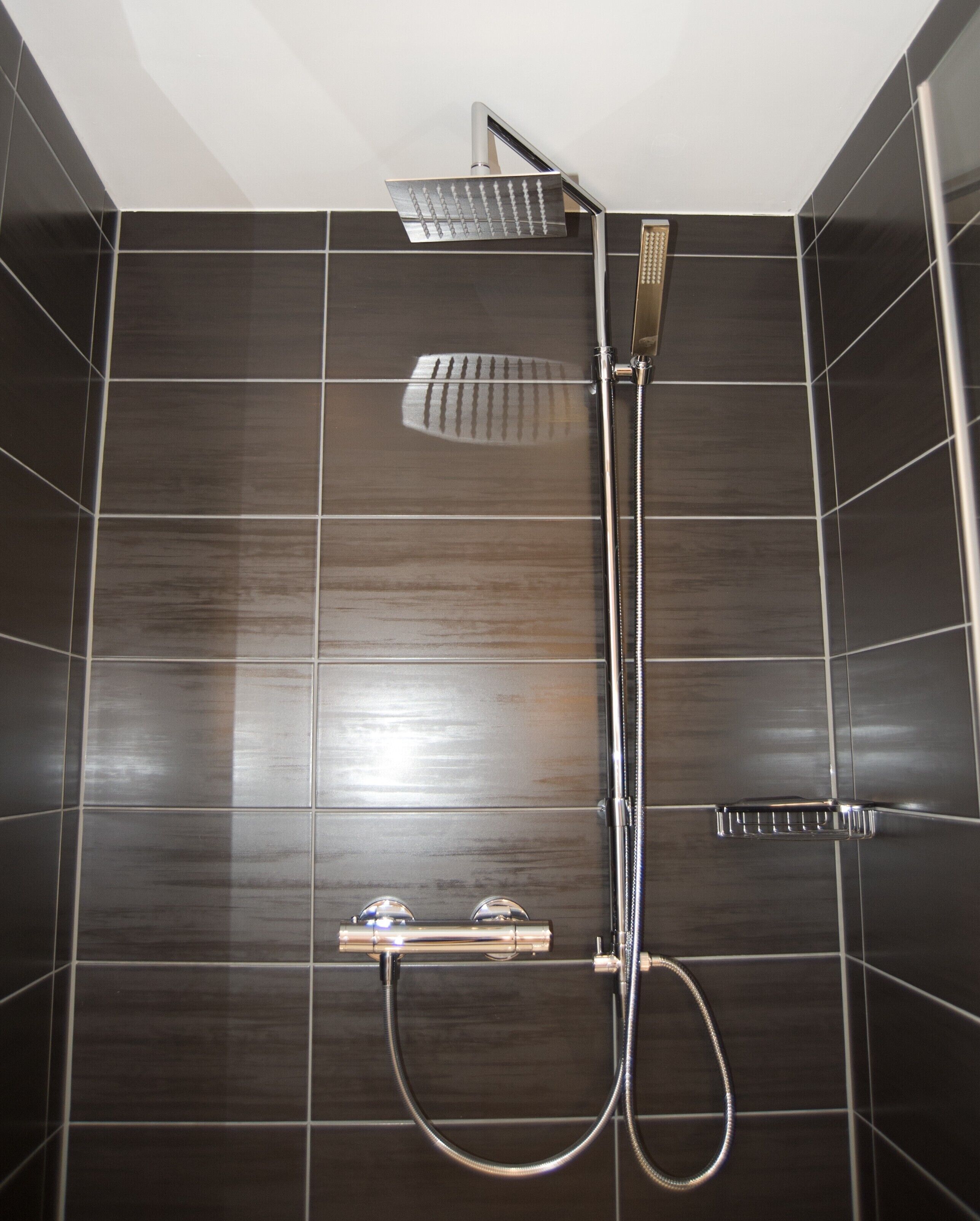 A shower with a handheld showerhead and a fixed rain showerhead, both mounted on a wall with dark tiles.
