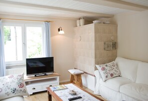 A room with a television on a wooden stand, a white sofa with floral cushions, a small wooden stool, and a table with a remote control and a book. - Holiday Home 'Kleines Landhaus Bodensee' with Lake View, Balcony & Wi-Fi (Überlingen)