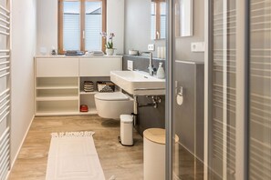 Un baño moderno con un tocador blanco, un inodoro, un bidé y una pequeña repisa con toallas y una planta.