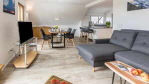 Un salon moderne avec un canapé gris, un téléviseur posé sur un meuble en verre, un coin repas avec une table et des chaises, et une cuisine en arrière-plan.