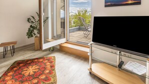 Un salon moderne doté d’un grand téléviseur, d’une porte-fenêtre coulissante donnant sur un balcon, d’un banc en bois et d’un tapis à motifs.