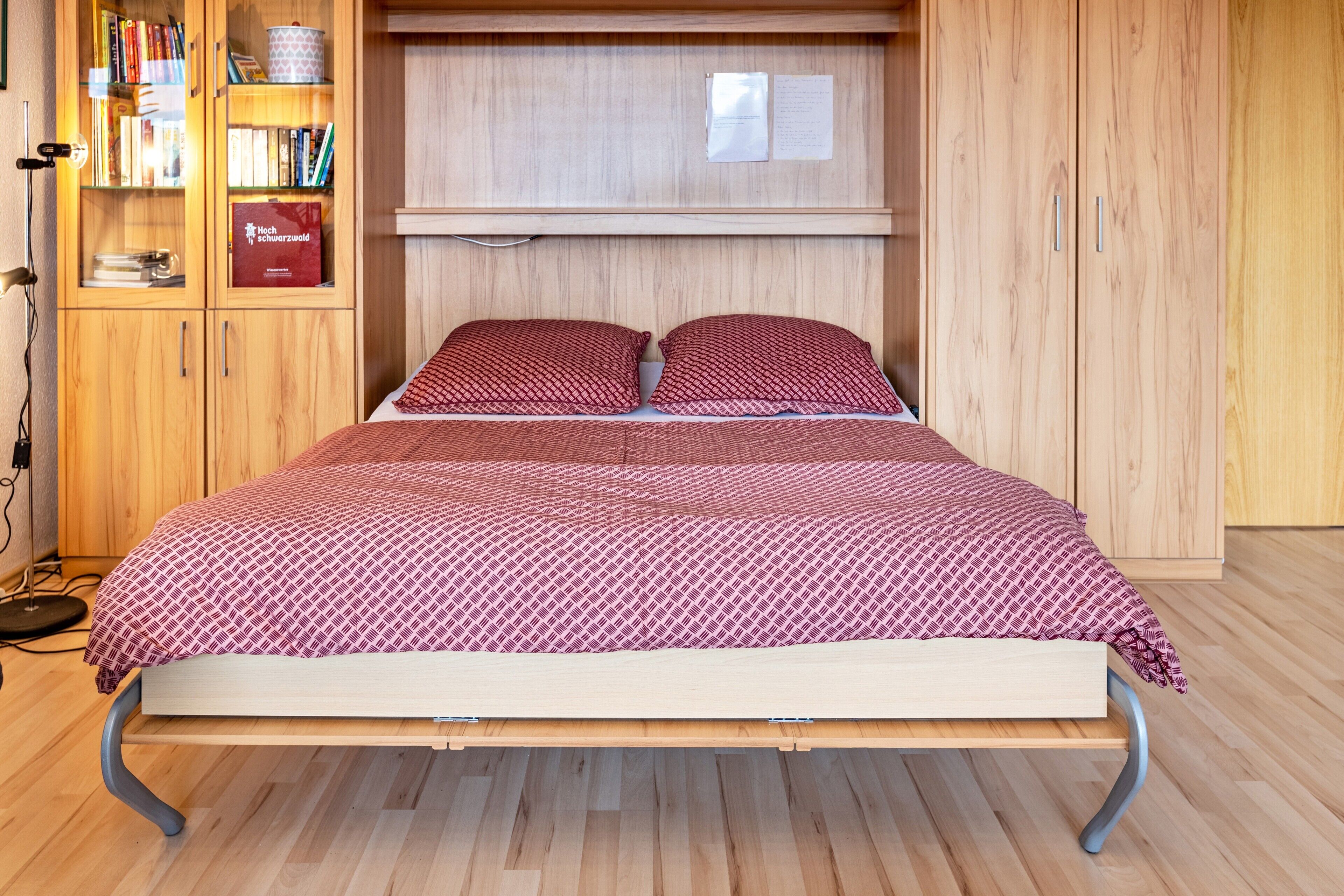 Una cama con una colcha estampada, un armario de madera con libros y una caja roja etiquetada 'Mech Schwerpunkt'.