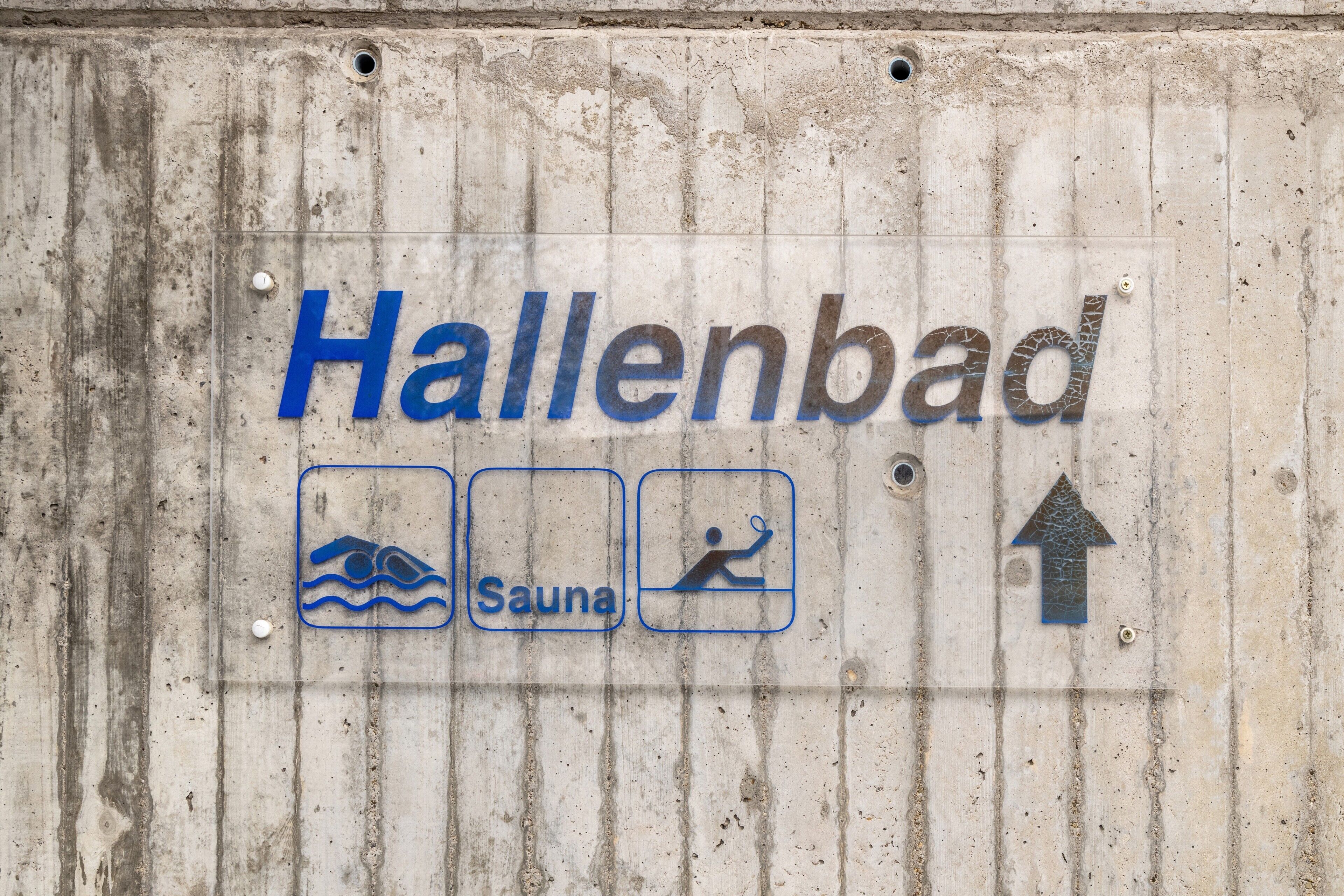 Segno con la scritta 'Hallenbad' e icone che rappresentano una piscina, una sauna e una freccia rivolta verso l'alto.