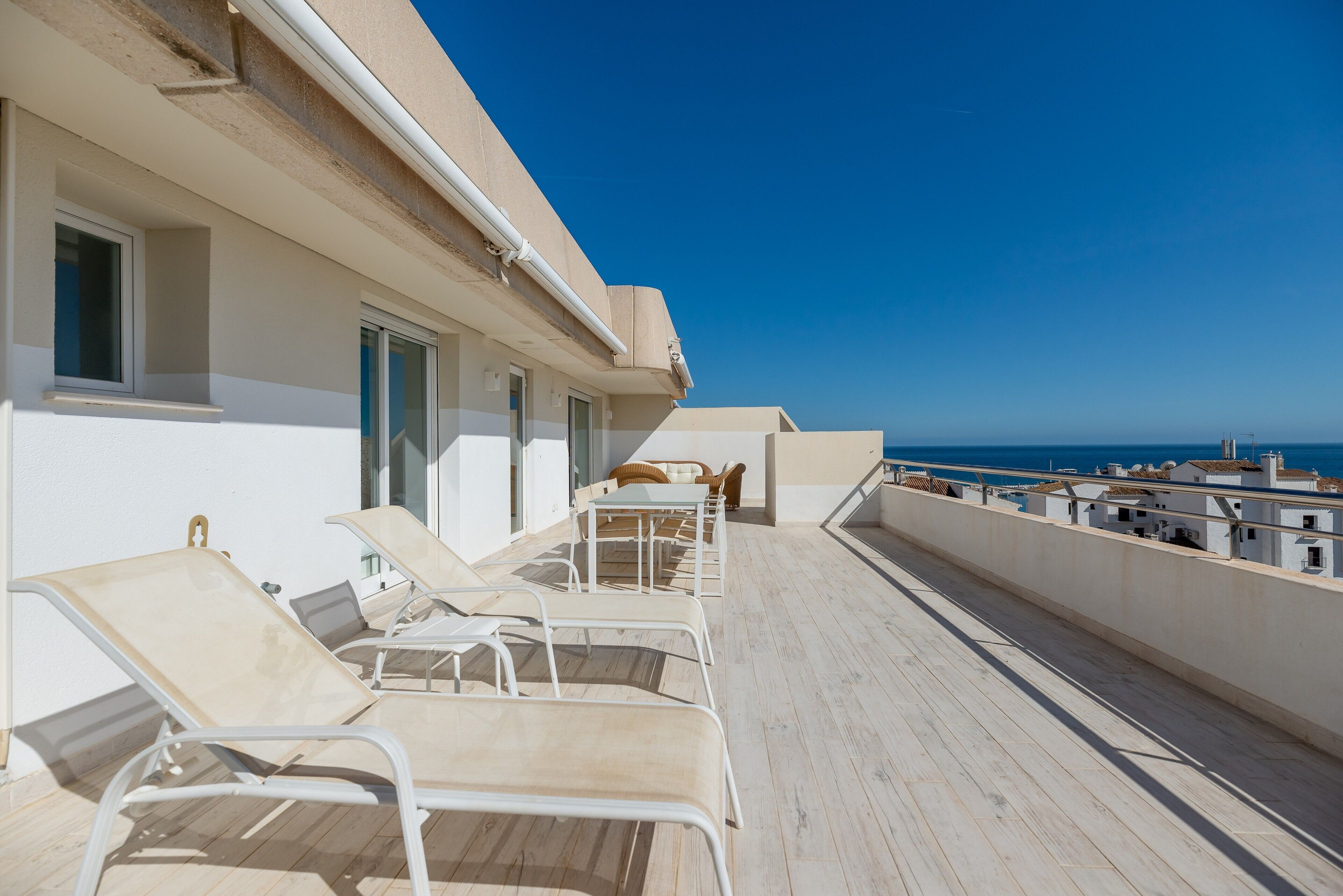 A balcony with sun loungers, a table, and chairs, overlooking a clear blue sky and a distant coastline.