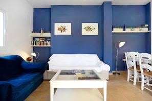 Un salon avec un canapé bleu, une table basse blanche et des chaises blanches. On y trouve des étagères remplies de livres et deux tableaux encadrés accrochés au mur.