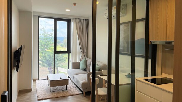 Ein modernes Wohnzimmer mit einer Couch, einem Couchtisch und einem Essbereich mit Stühlen und einem Tisch. Eine Glastür verschiebt sich zu einem Balkon mit Blick auf Grünflächen und ein Gebäude.