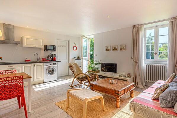 A modern kitchen with a washing machine, a dining area with a red chair, a sofa with a striped blanket, and a TV in the living room.