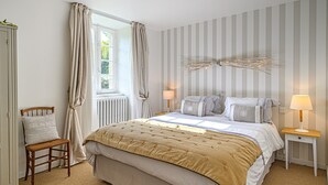 A bedroom with a bed, a chair, a nightstand, a lamp, and a window with curtains.