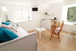 Une pièce lumineuse et blanche, avec un lit, une petite table, une chaise en osier, un coin cuisine et un téléviseur fixé au mur.