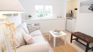 A living room with a sofa, a coffee table, a lamp, a bench, a window, and a kitchen area.