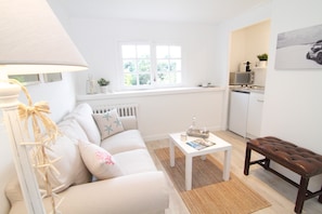 A living room with a sofa, a coffee table, a lamp, a bench, a window, and a kitchen area.