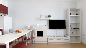 A modern kitchen with a dining table, chairs, a refrigerator, a TV, and a bookshelf.