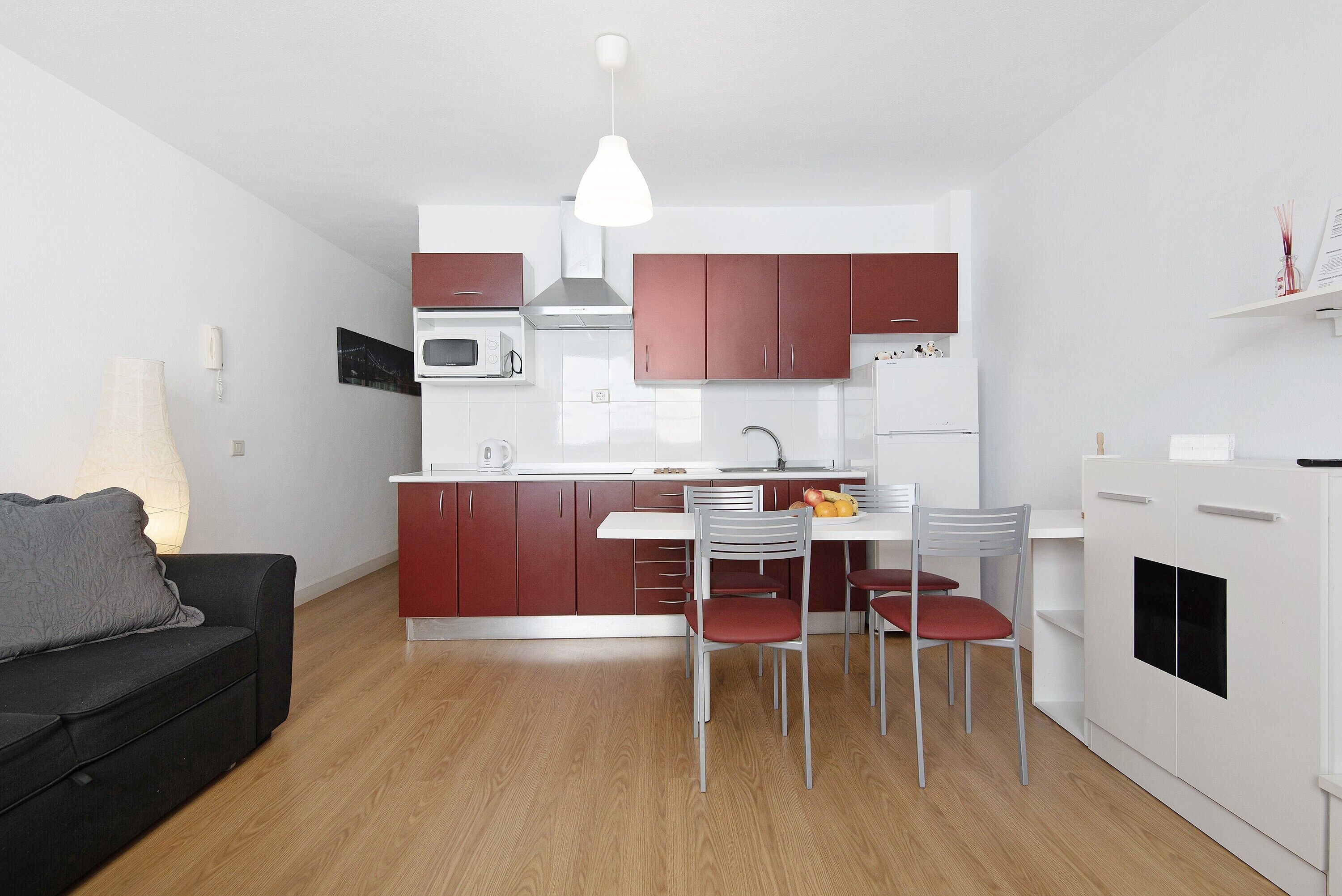 A modern kitchen with white cabinets, a dining table with chairs, a black sofa, and a pendant light.