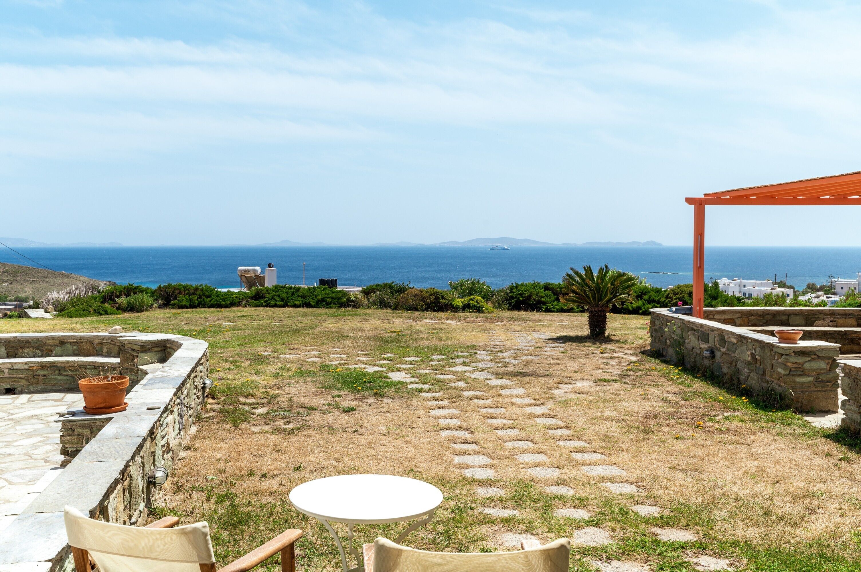 Eine Terrasse mit Steinfliesen, einem runden Tisch und Stühlen mit Blick auf die Küste, einschließlich einer Steinmauer und einer Pergola.