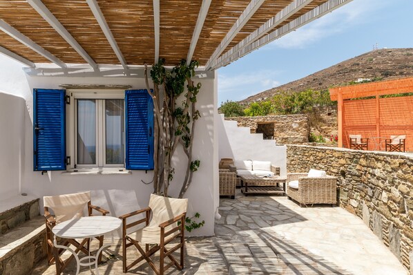 A covered outdoor seating area with stone walls, a white building, and a view of a hill.
