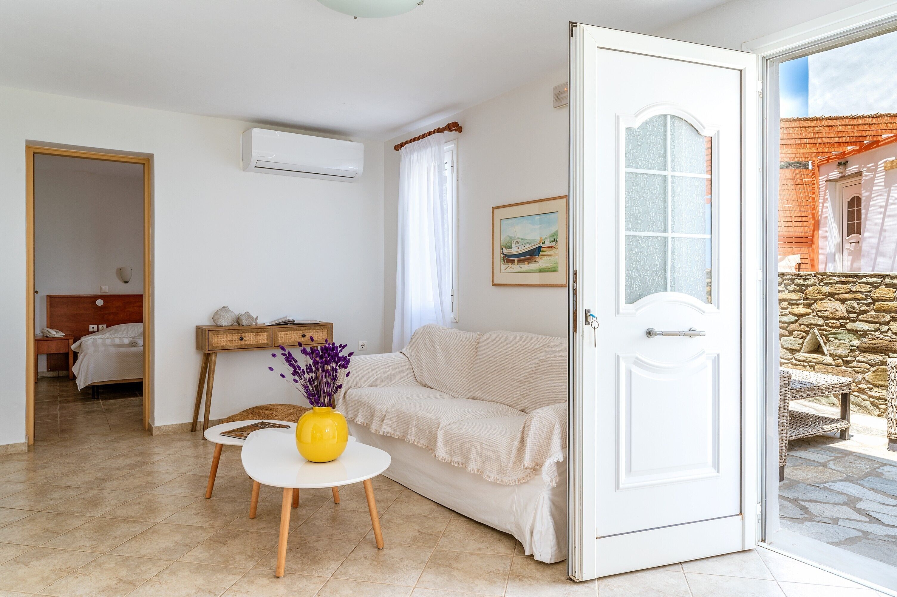 Een kamer met een witte bank, een klein tafeltje met een gele vaas, een houten bijzettafeltje, een bed op de achtergrond en een deur naar een buitenruimte met stenen muren.