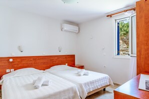 A hotel room with two beds, a wooden headboard, an air conditioning unit, a window with a view of greenery, and a wooden desk with a lamp.