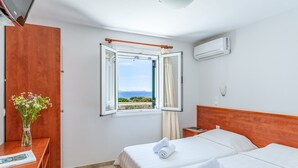A hotel room with two beds, a wooden wardrobe, a window with an open curtain, a vase with flowers, and an air conditioning unit. - Holiday Home 'Anemologio 101 Tinos' with Sea View, Wi-Fi and Air Conditioning (Laouti)