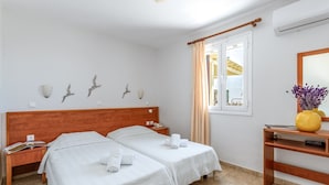 Habitación de hotel con dos camas, cabecero de madera, mesita de noche con teléfono, mesita de noche con una revista, ventana con cortinas, espejo y un jarrón con flores.