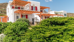 Un edificio blanco con contraventanas rojas, una pérgola de madera y abundante vegetación en primer plano.