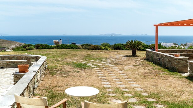 A patio with stone flooring, a round table, and chairs, overlooking a coastal view with a stone wall and a pergola. - Holiday Home 'Anemologio 204 Tinos' with Sea View, Wi-Fi and Air Conditioning (Laouti)