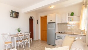 Cocina compacta con refrigerador, estufa y gabinetes blancos. Hay un comedor con mesa y sillas, y un sofá en la sala.