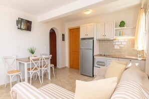Cocina compacta con refrigerador, estufa y gabinetes blancos. Hay un comedor con mesa y sillas, y un sofá en la sala.
