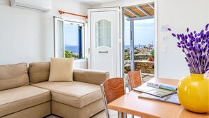 Una habitación con un sofá color beige, una mesa de madera, un jarrón amarillo con flores moradas y una puerta que da a un balcón con vistas.