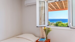 A bedroom with a bed, a nightstand with a magazine and a vase of flowers, and a window with a view of the sea.